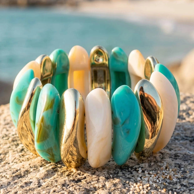 Turquoise marble and gold-tone resin cuff bracelet resting on a sandy rock with blurred sea in background