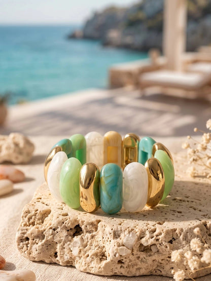 Gold-tone resin stretch bracelet with mint green, turquoise and white links displayed on a sandy limestone stone with ocean backdrop