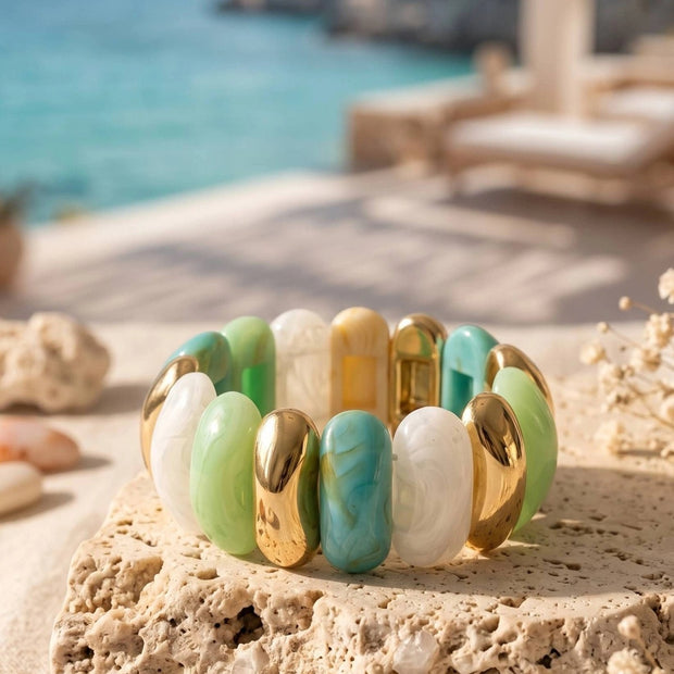 Gold-tone resin stretch bracelet with mint green, turquoise and white links displayed on a sandy limestone stone with ocean backdrop