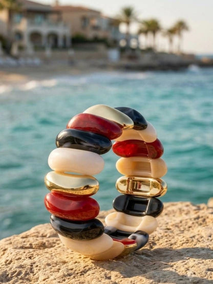 Multicolour resin cuff bracelet with red, black, ivory, and gold-tone oval tiles standing upright on a sandy rock by the sea