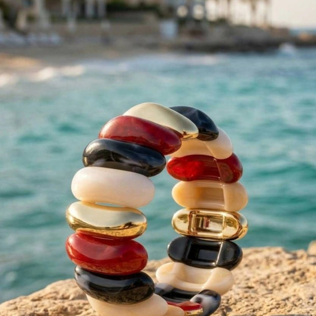 Multicolour resin cuff bracelet with red, black, ivory, and gold-tone oval tiles standing upright on a sandy rock by the sea