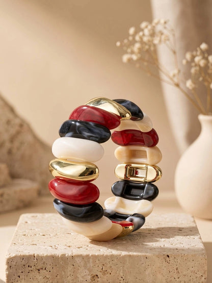 Multicolour resin cuff bracelet with red, black, ivory, and gold-tone tiles displayed upright on a travertine pedestal