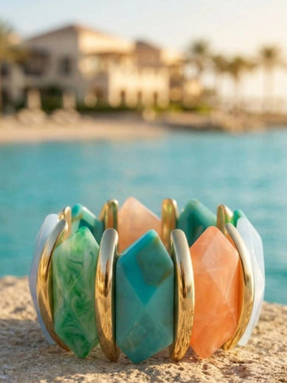 Multicolour resin stretch bracelet with gold-tone bars resting on sandy surface beside turquoise water