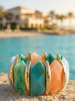 Multicolour resin stretch bracelet with gold-tone bars resting on sandy surface beside turquoise water