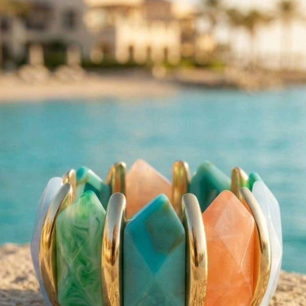 Multicolour resin stretch bracelet with gold-tone bars resting on sandy surface beside turquoise water