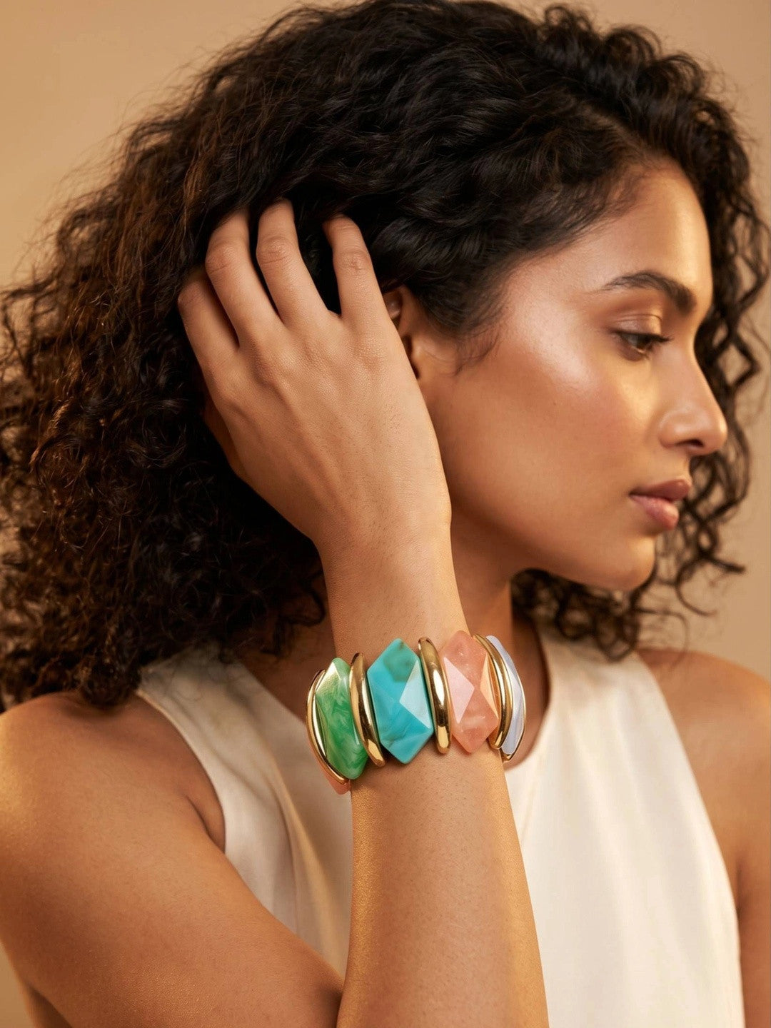 Model wearing multicolour resin stretch bracelet on wrist with hand raised near hair against warm backdrop