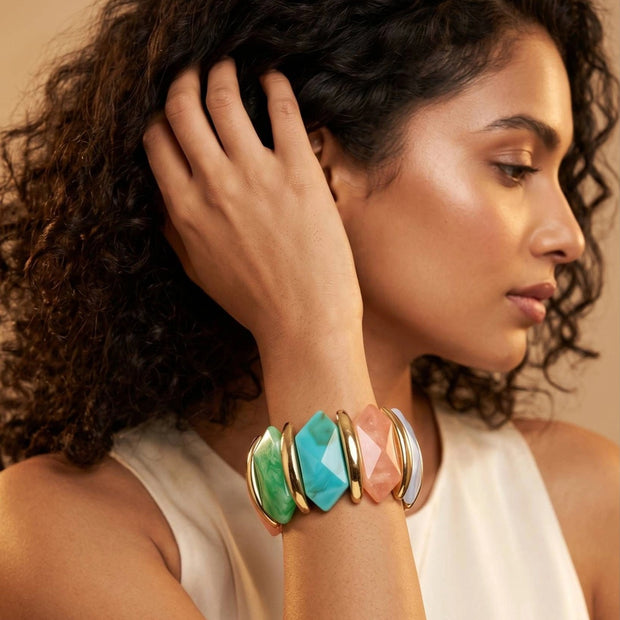 Model wearing multicolour resin stretch bracelet on wrist with hand raised near hair against warm backdrop