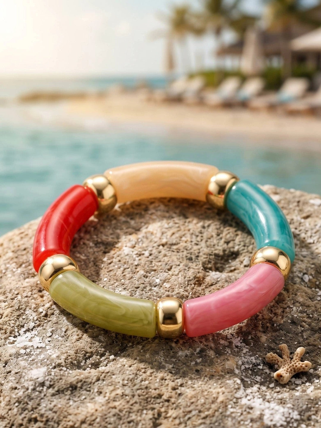 Multicolour resin tube stretch bracelet with gold-tone beads resting on a sandy rock by the sea
