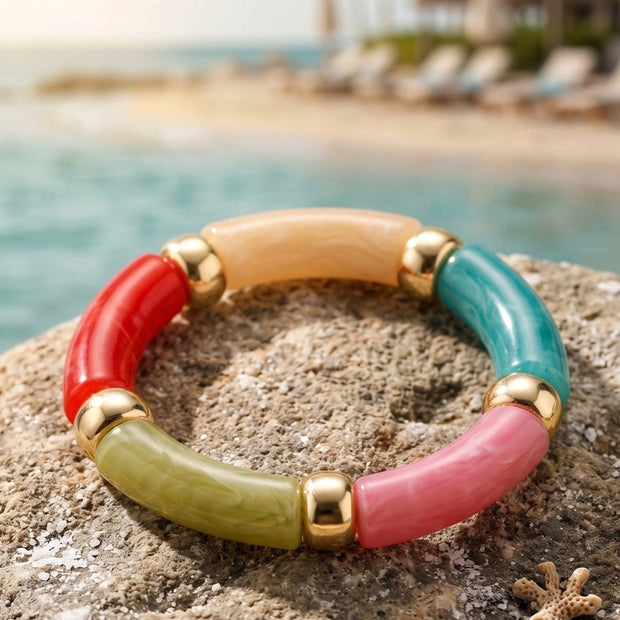 Multicolour resin tube stretch bracelet with gold-tone beads resting on a sandy rock by the sea