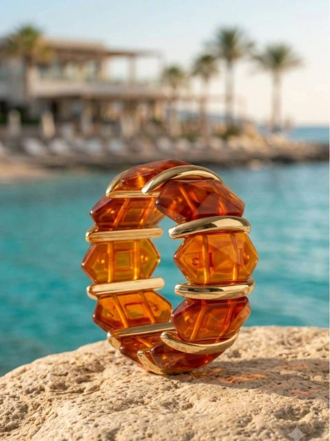 Amber resin and gold-toned cuff bracelet standing upright on a rocky surface beside turquoise sea