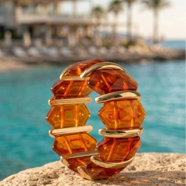 Amber resin and gold-toned cuff bracelet standing upright on a rocky surface beside turquoise sea