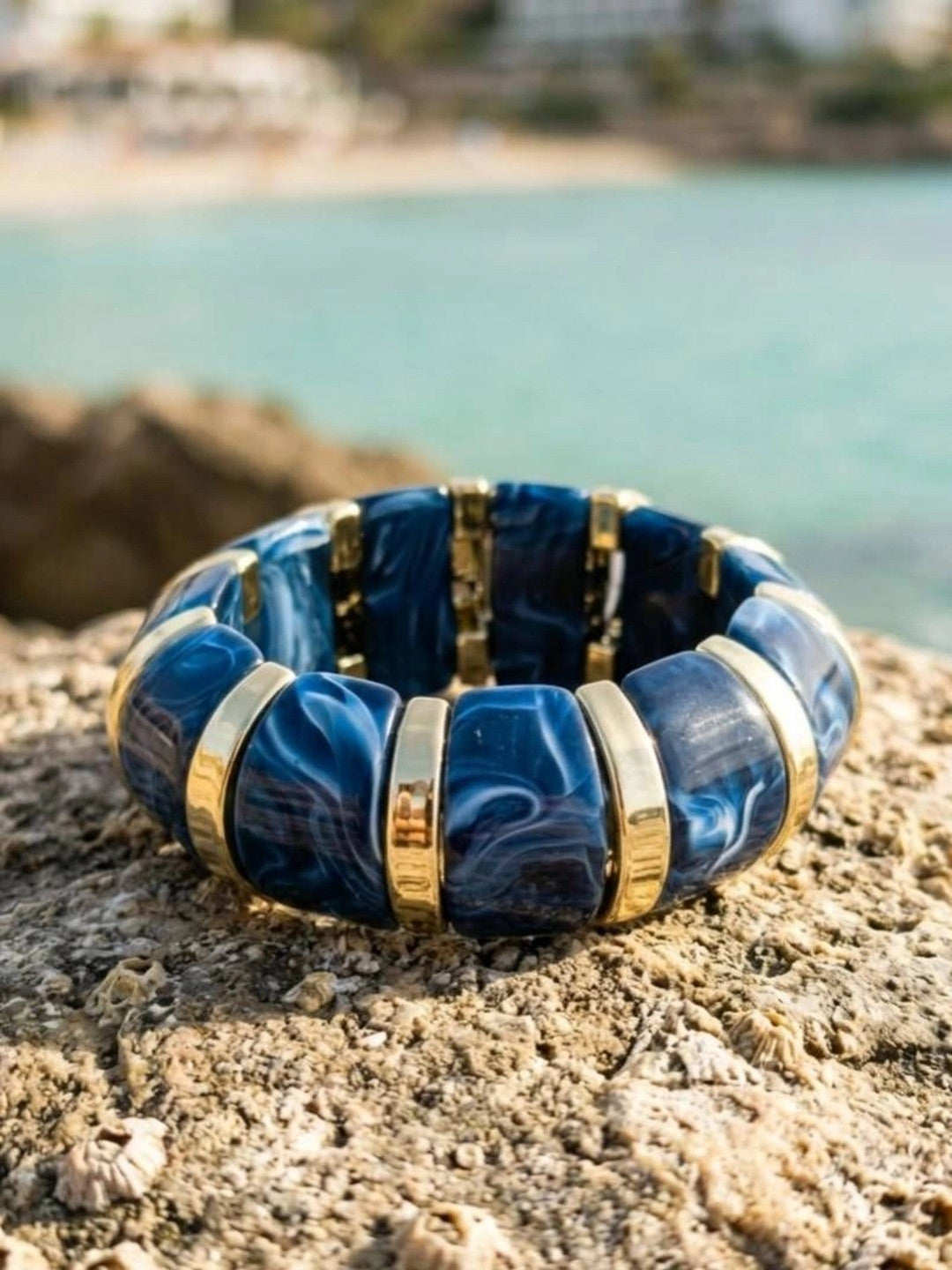 Navy blue marble resin stretch bracelet with gold-tone spacers resting flat on a rocky coastal surface with turquoise sea in background
