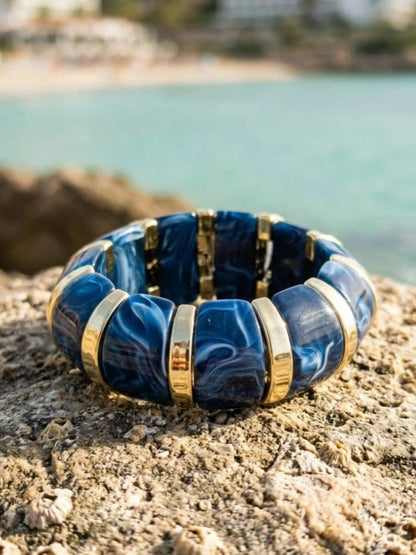 Navy blue marble resin stretch bracelet with gold-tone spacers resting flat on a rocky coastal surface with turquoise sea in background