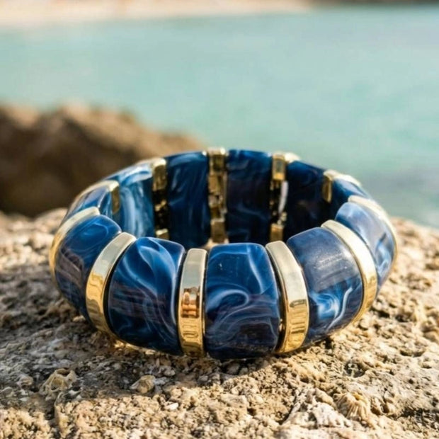 Navy blue marble resin stretch bracelet with gold-tone spacers resting flat on a rocky coastal surface with turquoise sea in background