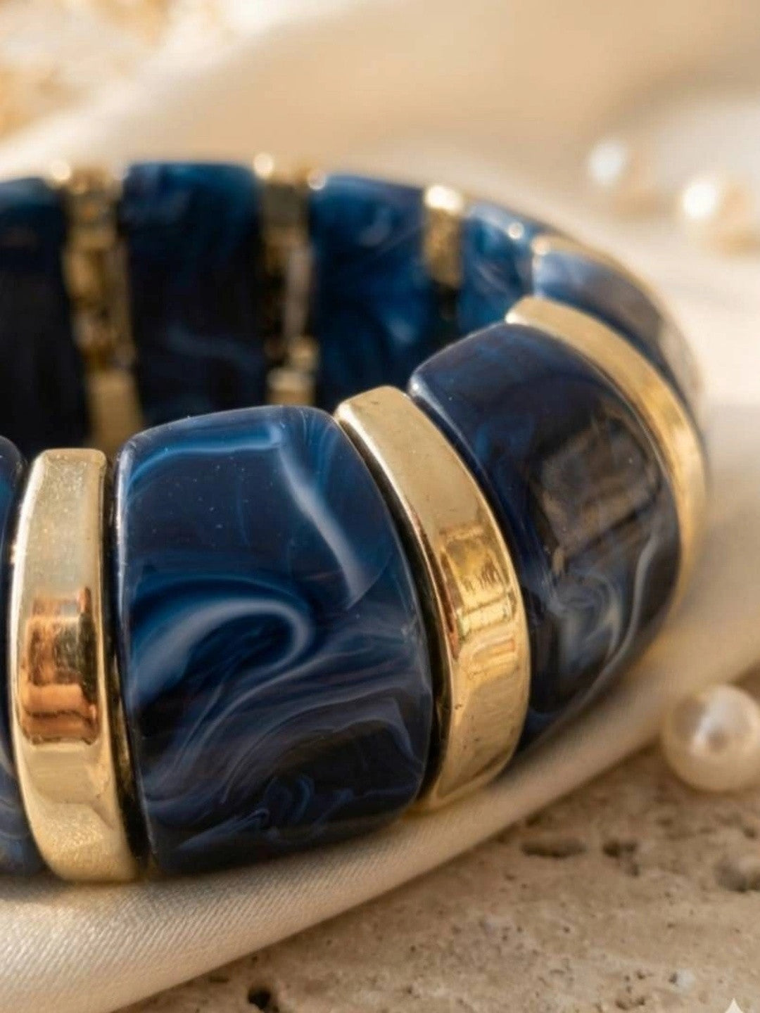Close-up of navy blue swirled resin tiles and polished gold-tone spacers on a stretch bracelet, styled with loose pearls on fabric