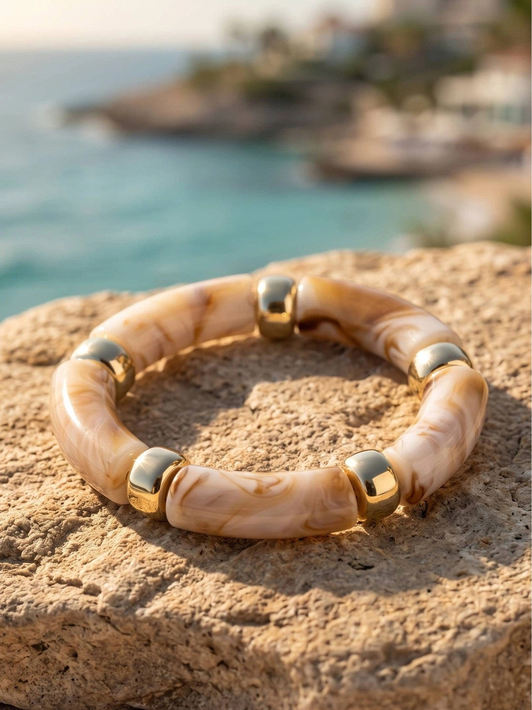 Cream marble resin bracelet with polished gold-tone spacers resting on a sandy rock beside turquoise water