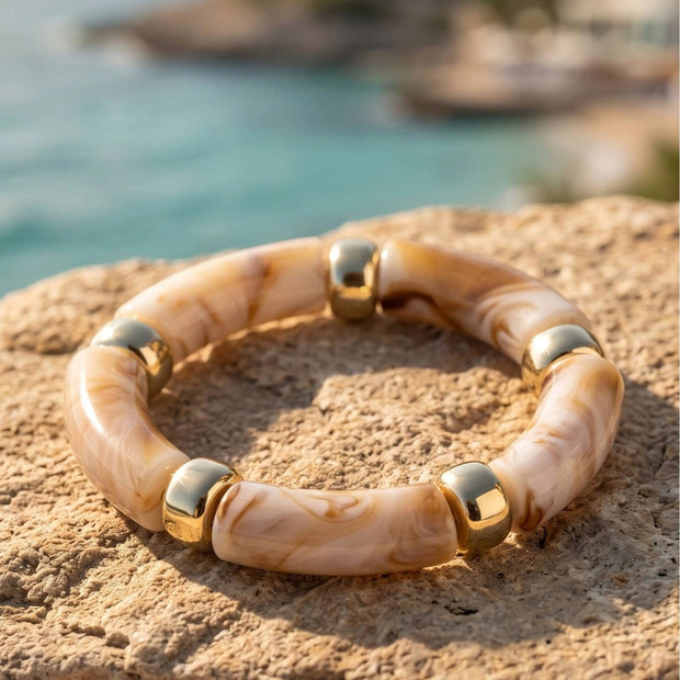 Cream marble resin bracelet with polished gold-tone spacers resting on a sandy rock beside turquoise water