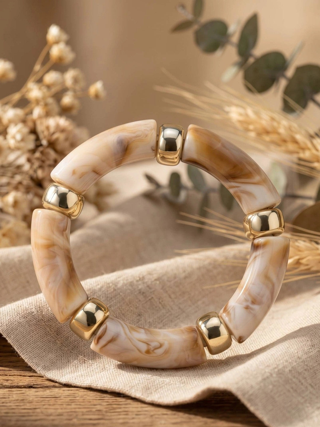 Cream and caramel marble resin bracelet with gold-tone beads laid on linen cloth with dried botanicals
