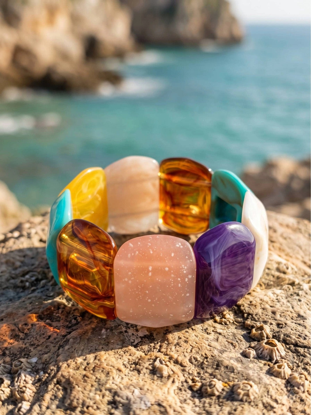 Multicolour resin stretch bracelet with chunky amber, rose, violet and teal stones resting on a coastal rock with turquoise sea in background