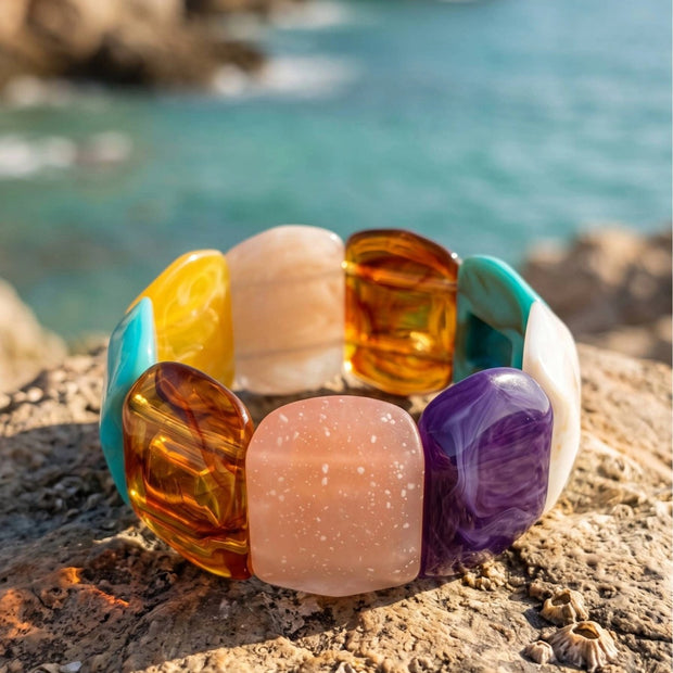 Multicolour resin stretch bracelet with chunky amber, rose, violet and teal stones resting on a coastal rock with turquoise sea in background