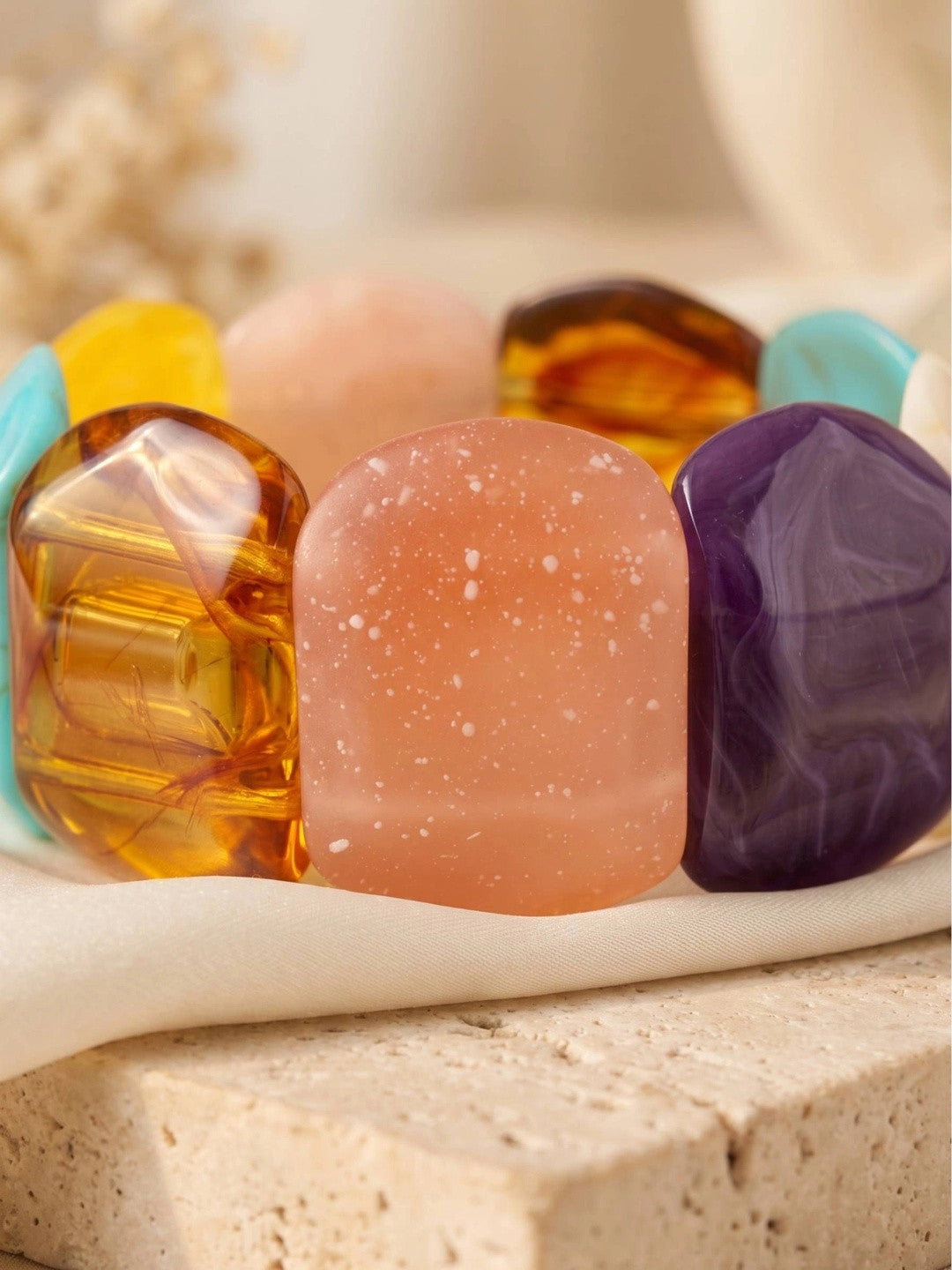 Close-up of multicolour resin bracelet showing translucent amber, frosted rose and marbled violet stones on a cream fabric surface