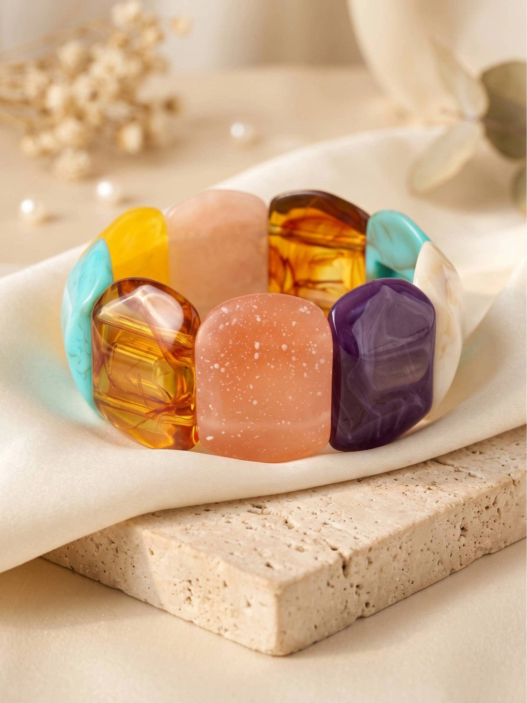 Multicolour chunky resin stretch bracelet laid flat on white silk cloth atop a travertine block with dried flowers in background
