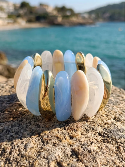 Blue marble cream and gold-tone stretch bracelet resting on a rocky surface with turquoise sea in background