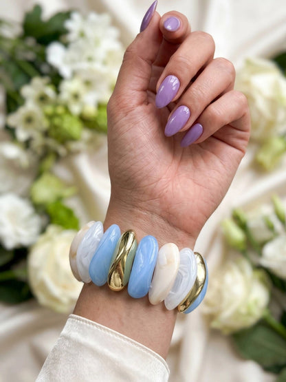Blue marble and gold-tone stretch bracelet worn on wrist of woman with lavender nail polish against floral background