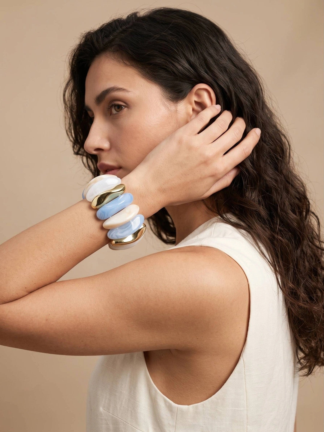 Woman in white tank top wearing blue marble and gold-tone stretch bracelet with hand raised near face