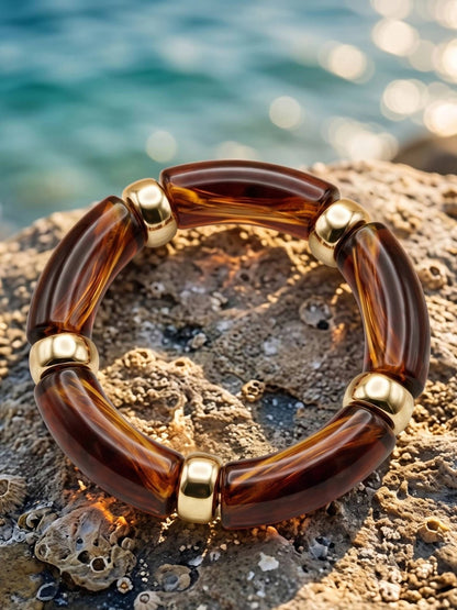 Tortoise resin stretch bracelet with gold-tone spacers resting flat on a textured coastal rock with turquoise water in the background