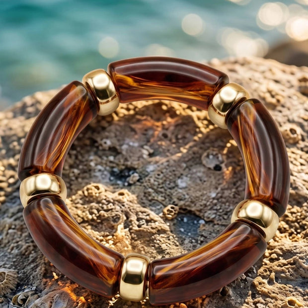 Tortoise resin stretch bracelet with gold-tone spacers resting flat on a textured coastal rock with turquoise water in the background