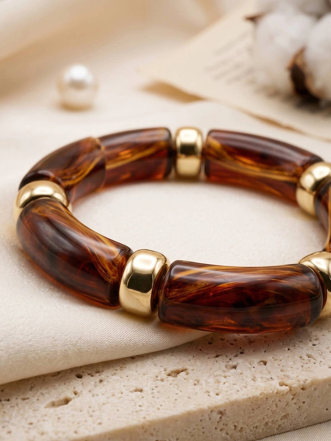 Close-up of amber tortoise resin bracelet with polished gold-tone metal beads laid on cream fabric beside a pearl and cotton stems