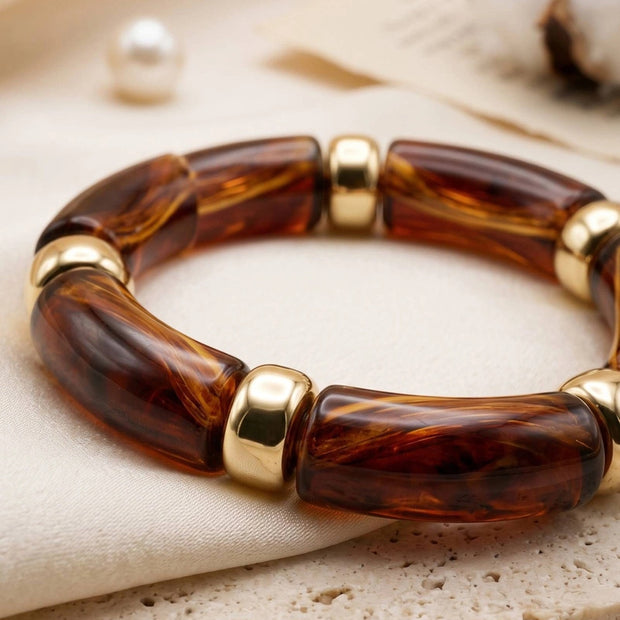 Close-up of amber tortoise resin bracelet with polished gold-tone metal beads laid on cream fabric beside a pearl and cotton stems