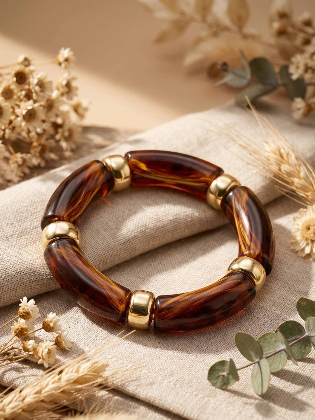 Tortoise resin gold-tone stretch bracelet placed on linen cloth surrounded by dried wildflowers and eucalyptus sprigs