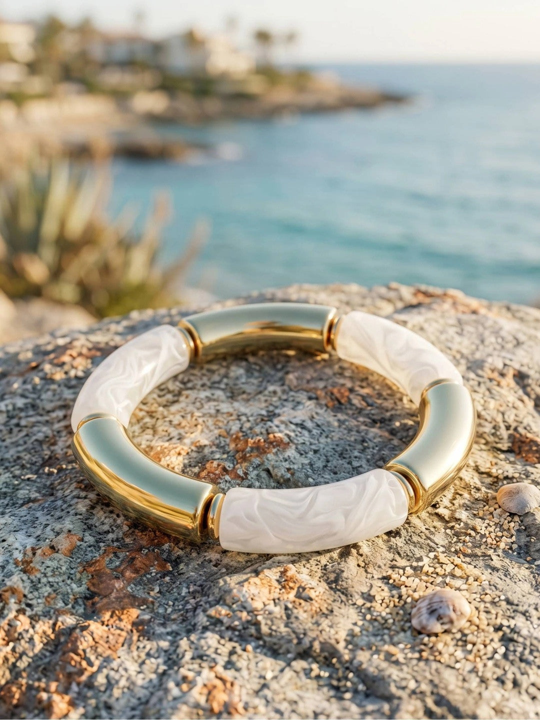 Gold-plated white resin tube stretch bracelet laid flat on a coastal rock with blue sea in the background