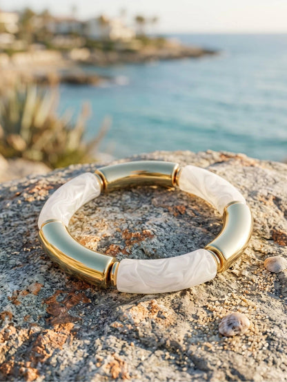 Gold-plated white resin tube stretch bracelet laid flat on a coastal rock with blue sea in the background