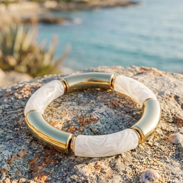 Gold-plated white resin tube stretch bracelet laid flat on a coastal rock with blue sea in the background