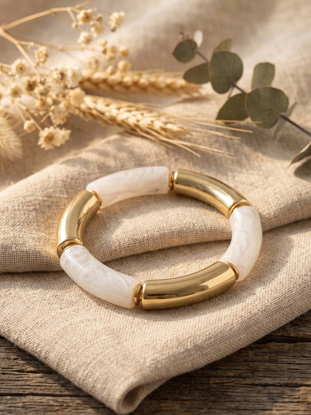 Gold-plated white resin stretch bracelet resting on folded linen cloth with dried botanicals in the background