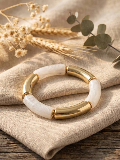 Gold-plated white resin stretch bracelet resting on folded linen cloth with dried botanicals in the background