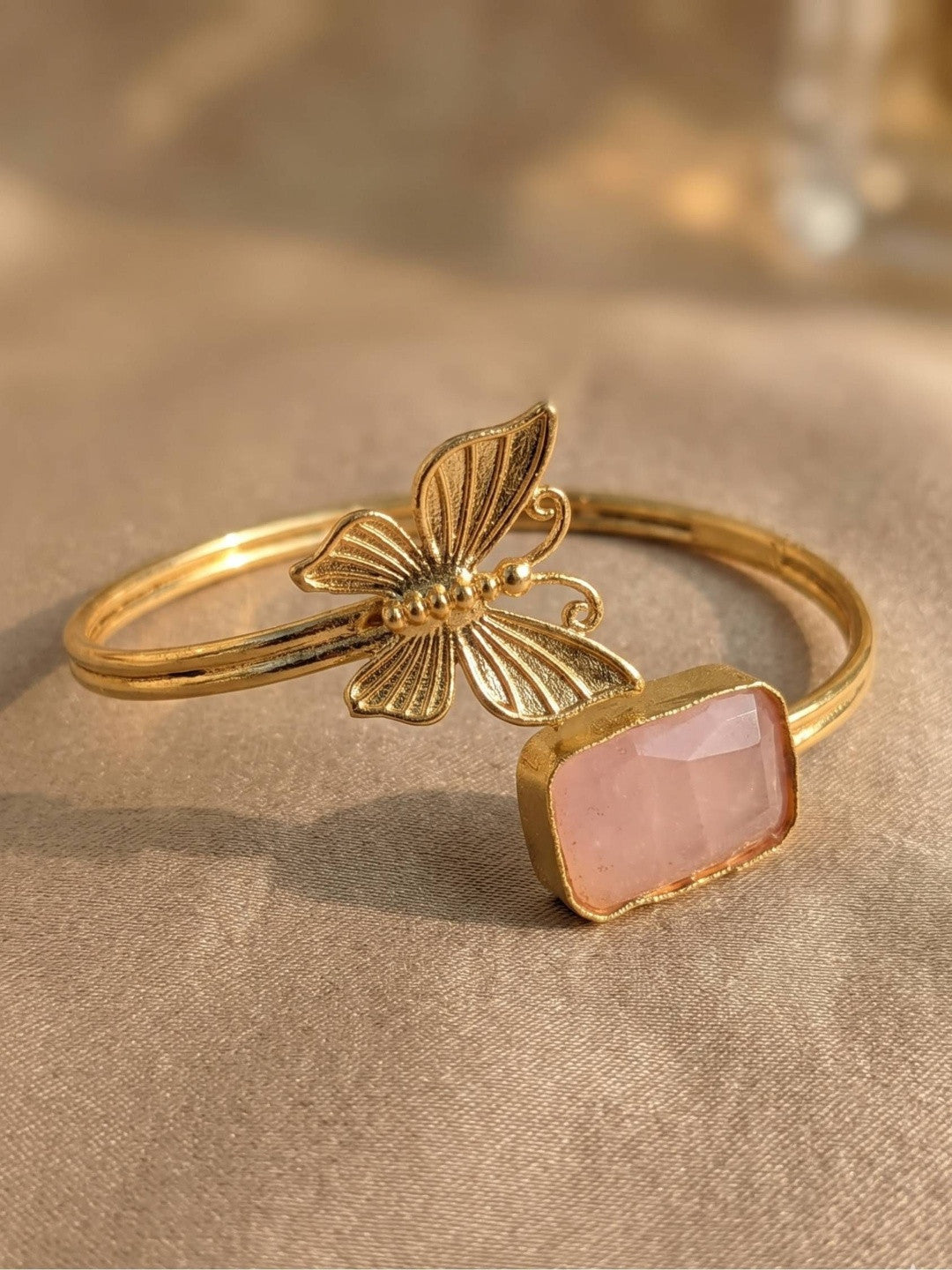 Gold-plated brass butterfly cuff bracelet with pink rectangular cabochon stone on a beige fabric surface