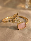 Gold-plated brass butterfly cuff bracelet with pink rectangular cabochon stone on a beige fabric surface