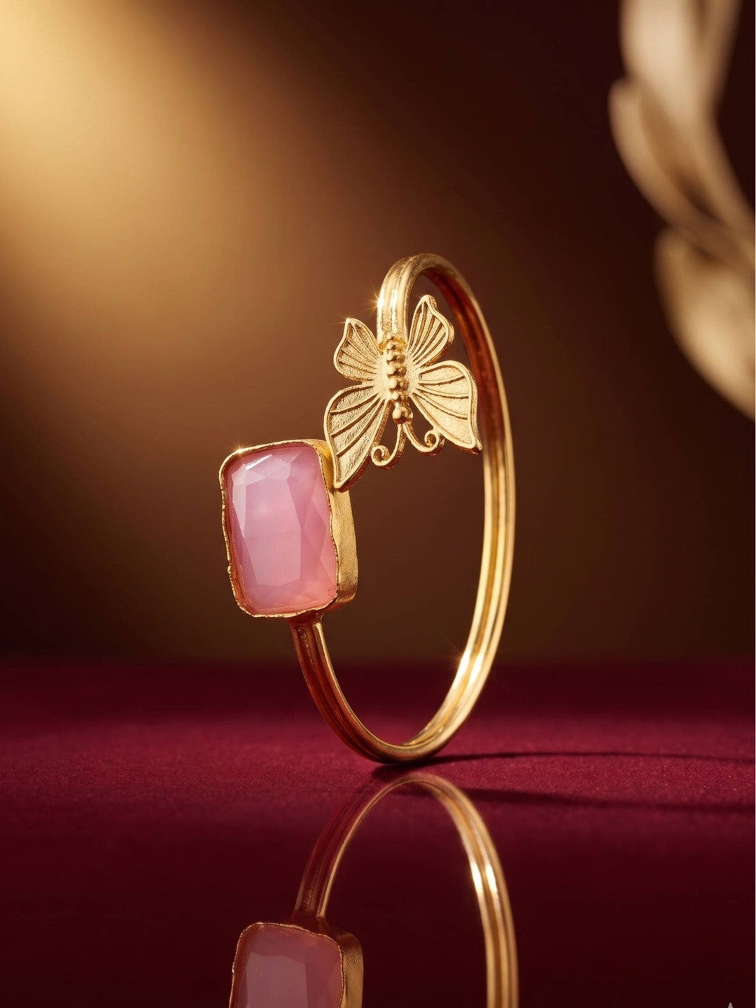 Side profile of gold butterfly cuff bracelet standing upright on a deep red reflective surface with warm lighting