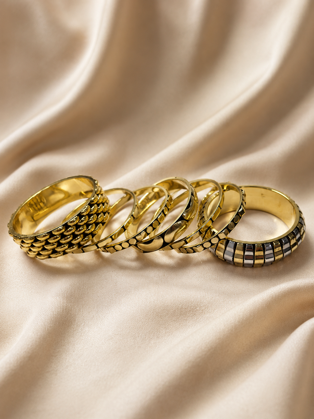 Five gold-tone textured bangles laid flat on cream satin fabric showing varied surface patterns