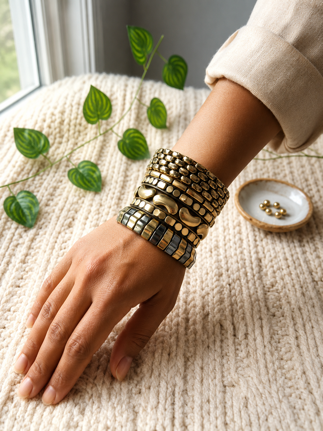 Hand resting on a knit surface wearing a full stack of five gold textured bangles near green leaves