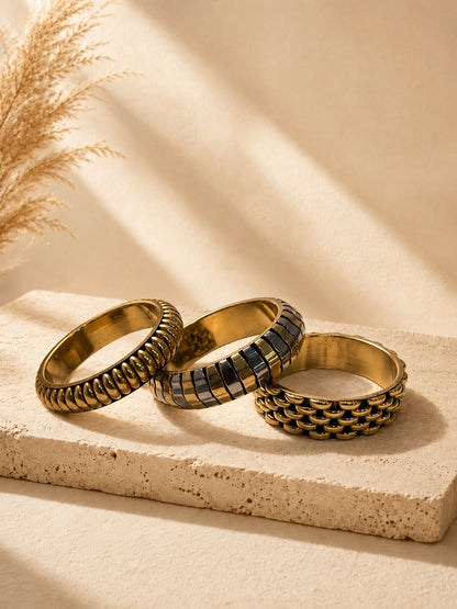 Three gold-tone textured metal bangles laid flat on a travertine surface with pampas grass in the background