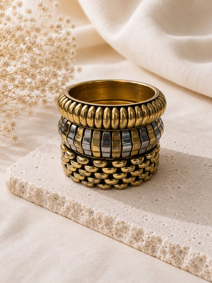 Three stacked gold-tone bangles showing ribbed, two-tone striped and fish-scale textures on a stone platform with dried flowers