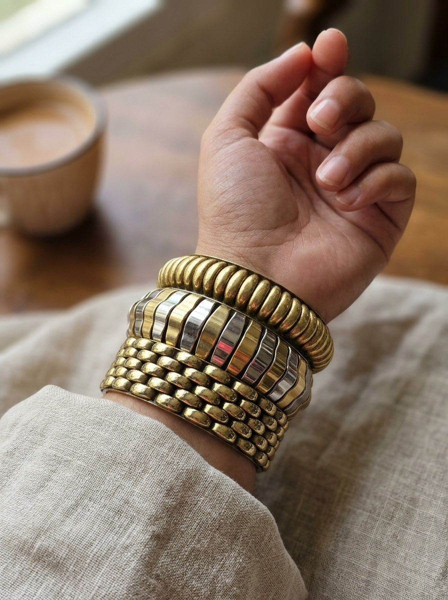 All three gold-tone textured bangles stacked and worn on a wrist against a linen sleeve with a coffee cup in the background