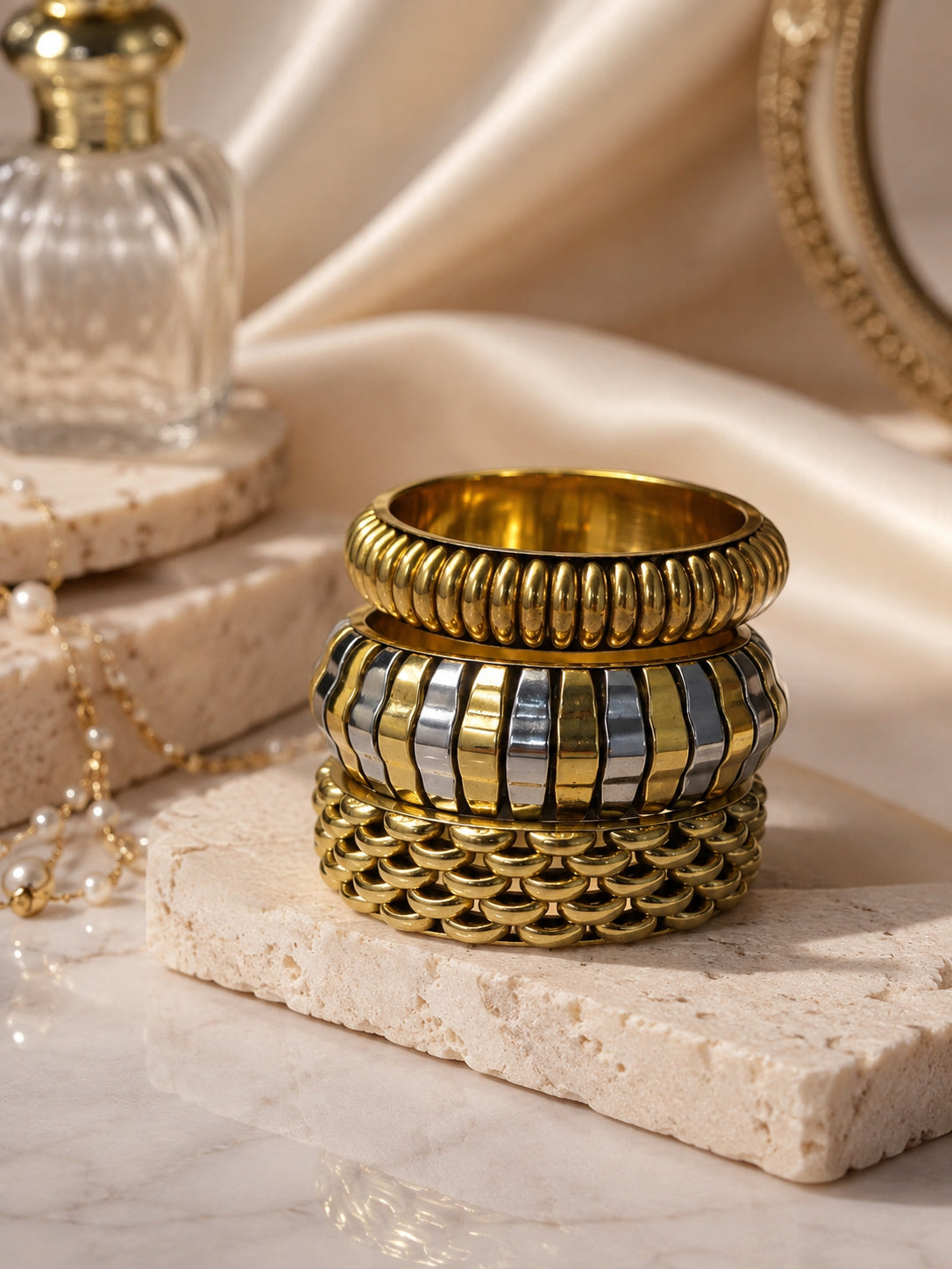 Gold-tone textured bangle stack on travertine stone styled with a perfume bottle, pearl necklace and silk fabric backdrop