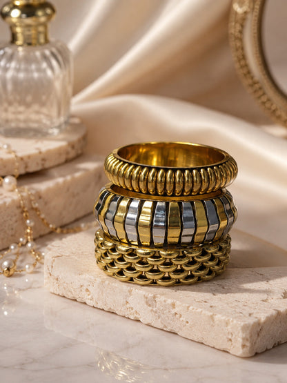 Gold-tone textured bangle stack on travertine stone styled with a perfume bottle, pearl necklace and silk fabric backdrop