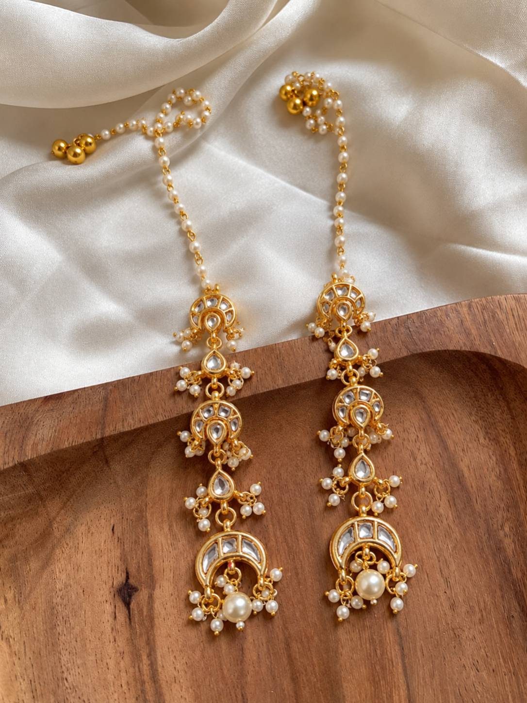Two kundan chandbali earrings with pearl drops laid flat on a wooden tray with white fabric
